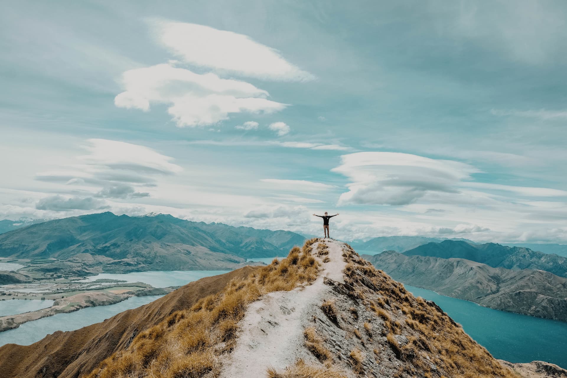 A person standing on a mountain ridge with arms outstretched over a wide valley and lake